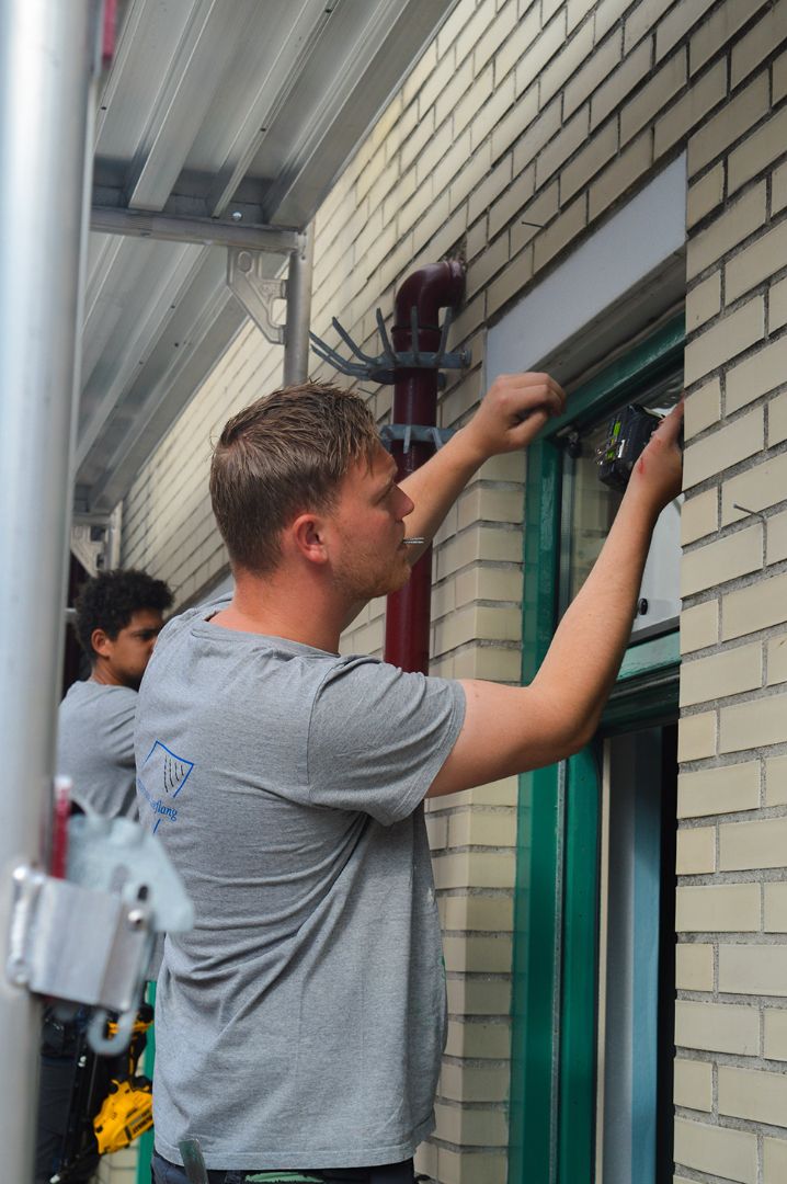 Deze afbeelding toont twee mannen die werken aan een raam van een gebouw. De man op de voorgrond draagt een grijs T-shirt met het logo en de tekst "Glasservice Leeflang" op de rug. Hij staat met zijn handen in zijn zij en kijkt omhoog.